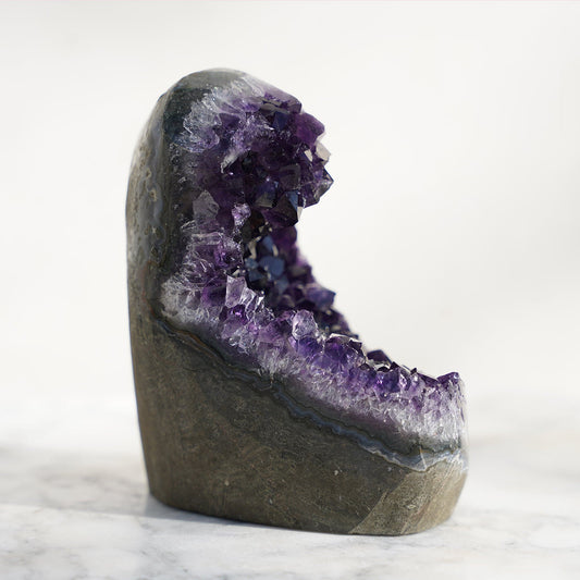 Cut-base cluster style amethyst in the shape of an ocean's wave. Traditional polish displays delightful bands of layered minerals including white quartz, light blue agate, and green celadonite outlining the raw basalt rock in the back. 