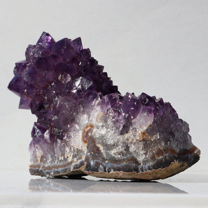 Amethyst cluster in the shape of a nature-made medieval boot. In plum color crystal peaks with a significant portion of the quartz crystals in white, bands of orange and blue agate.